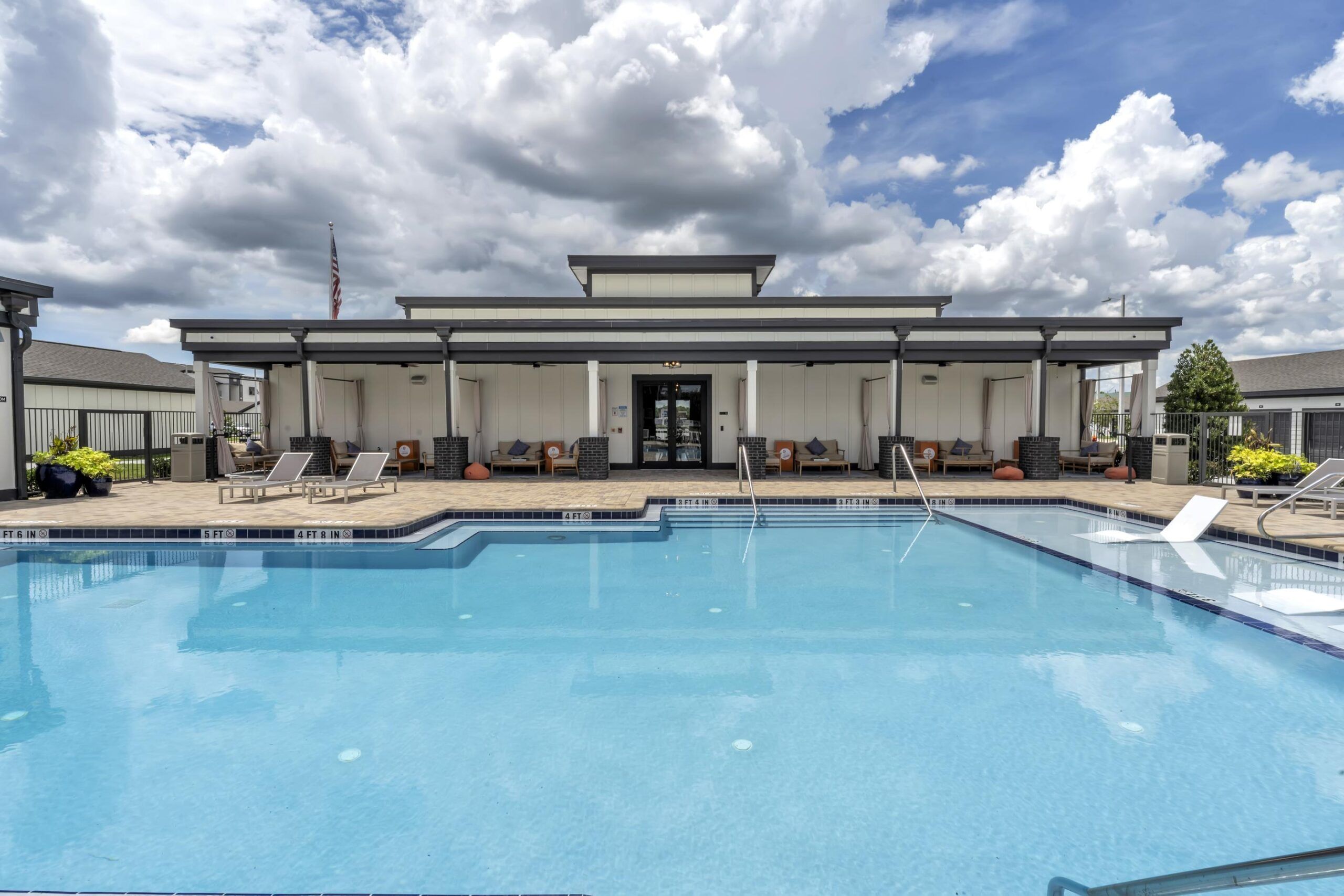 Outdoor swimming pool with lounge chairs, umbrellas, and a modern building in the background under a blue sky.