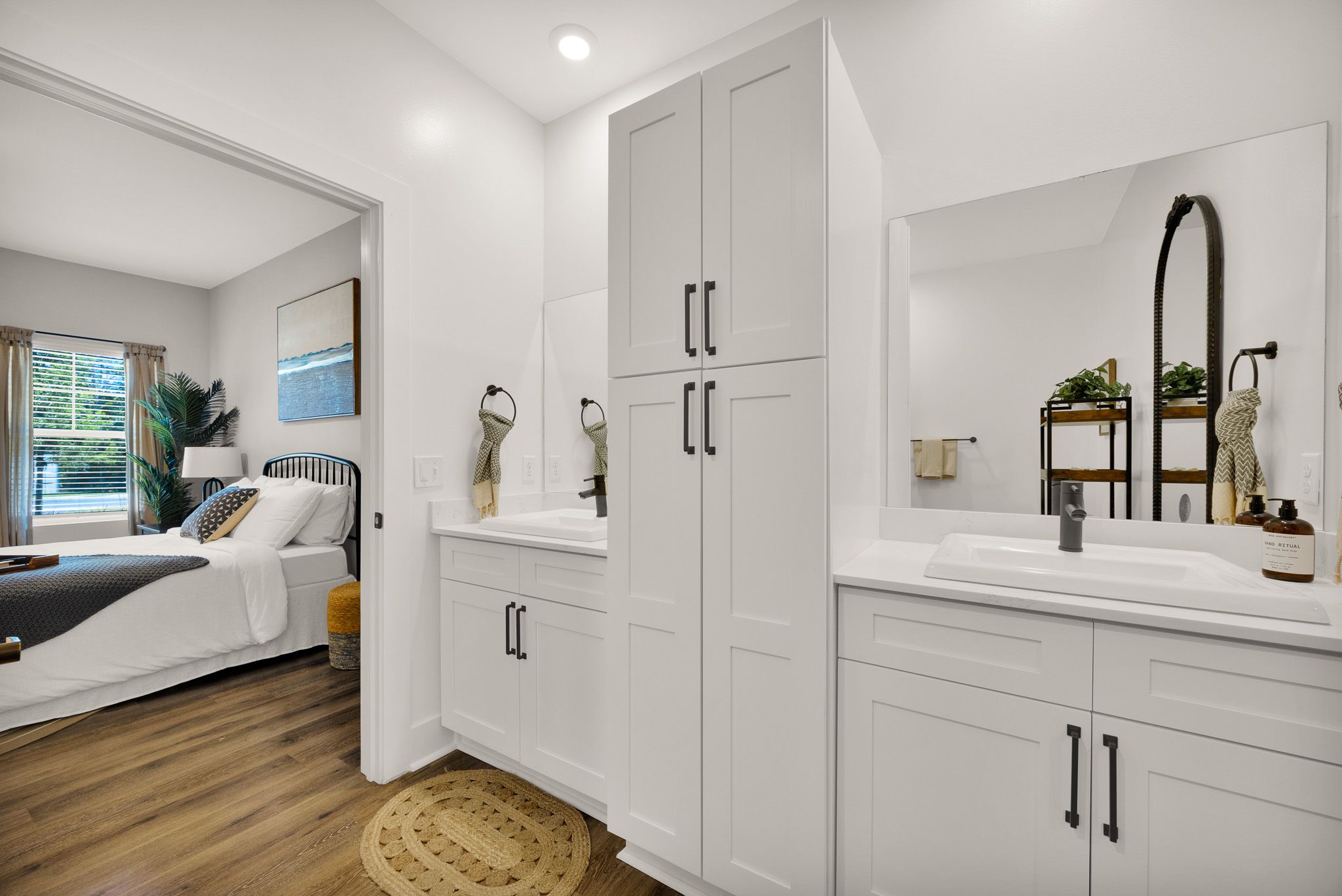 Modern bathroom with double sinks, white cabinets, and a view into a cozy bedroom with a large window.