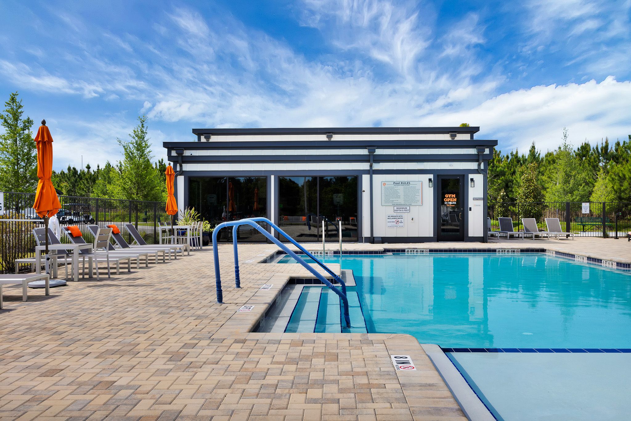 Outdoor swimming pool with lounge chairs, umbrellas, and a modern building in the background under a blue sky.