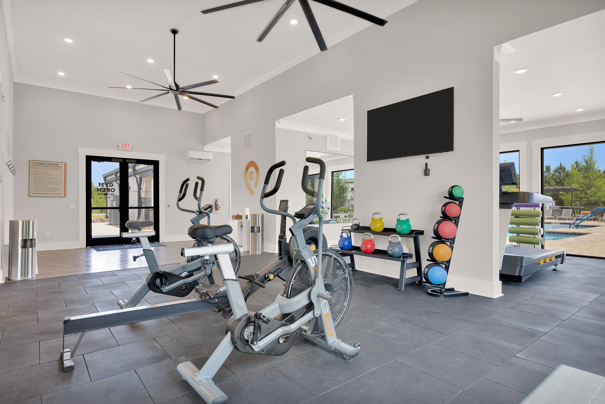 Modern gym room with two stationary bikes, free weights, TV, and large windows overlooking a pool.
