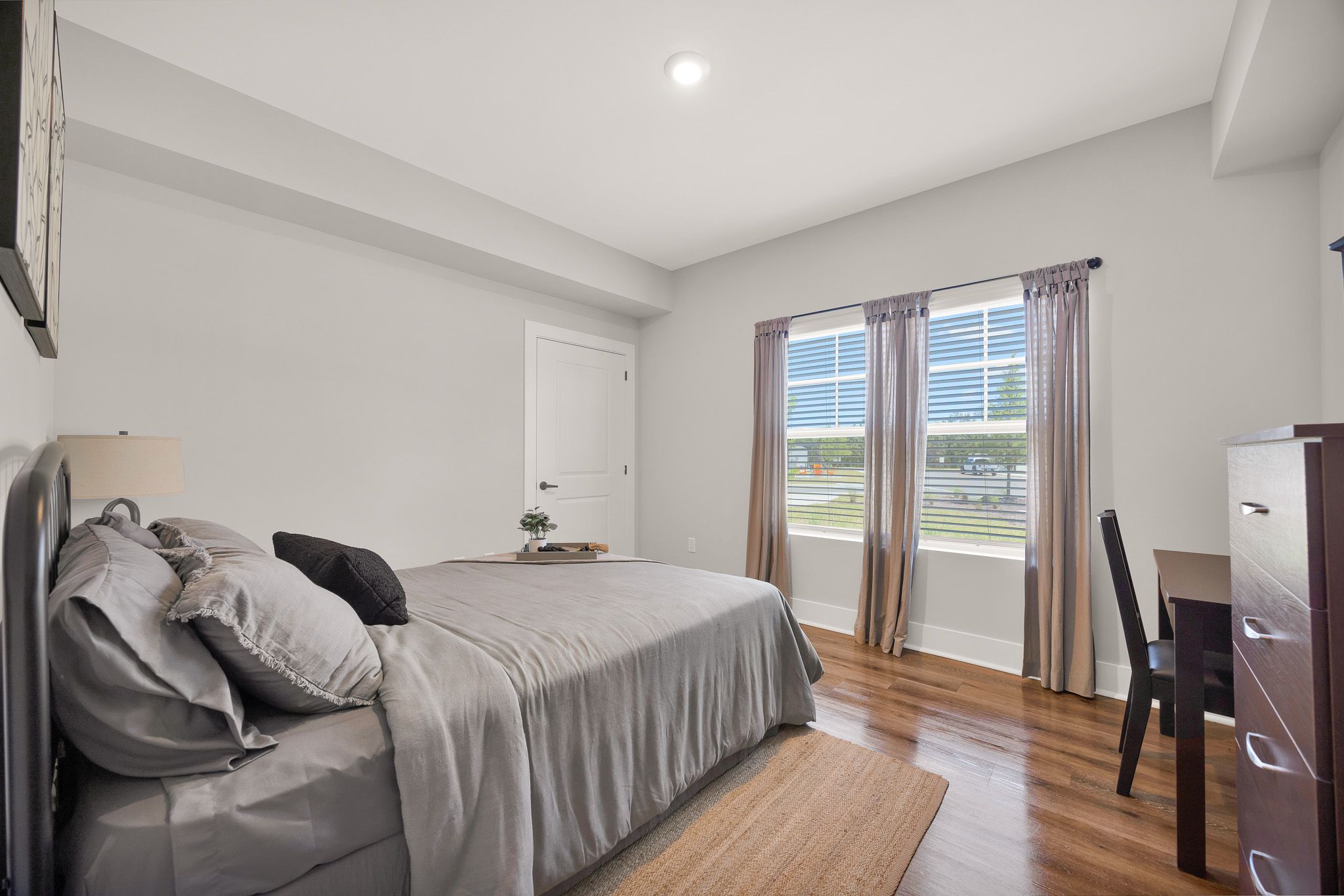 Bright bedroom with a neatly made bed, wooden floors, a desk by the window, and neutral-colored decor.