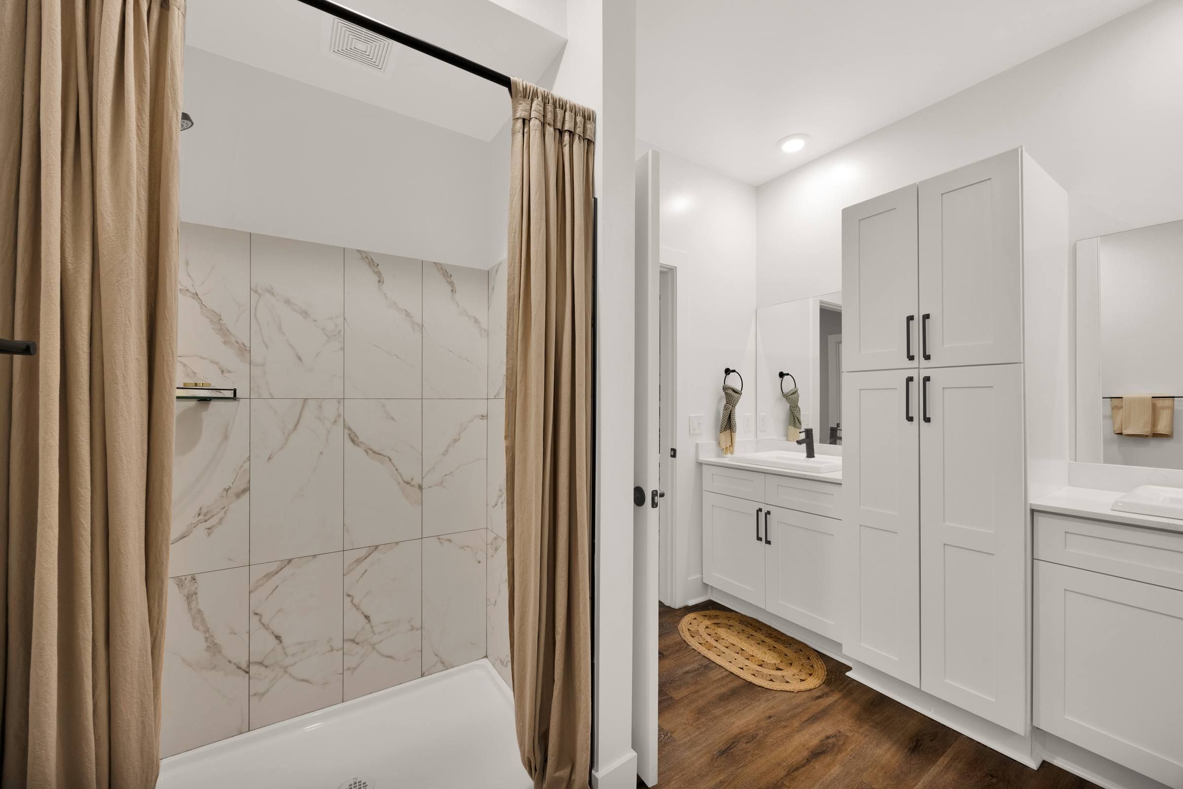 Modern bathroom with a tiled shower, beige curtains, white cabinets, and a wood floor.