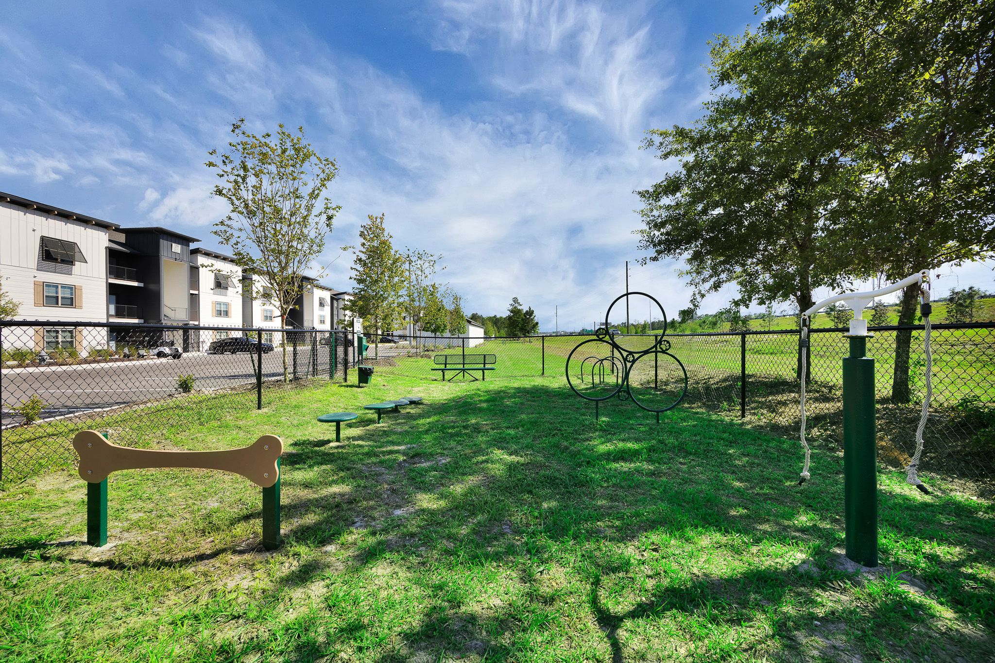 A fenced dog park with agility equipment, green grass, trees, and apartments in the background under a blue sky.