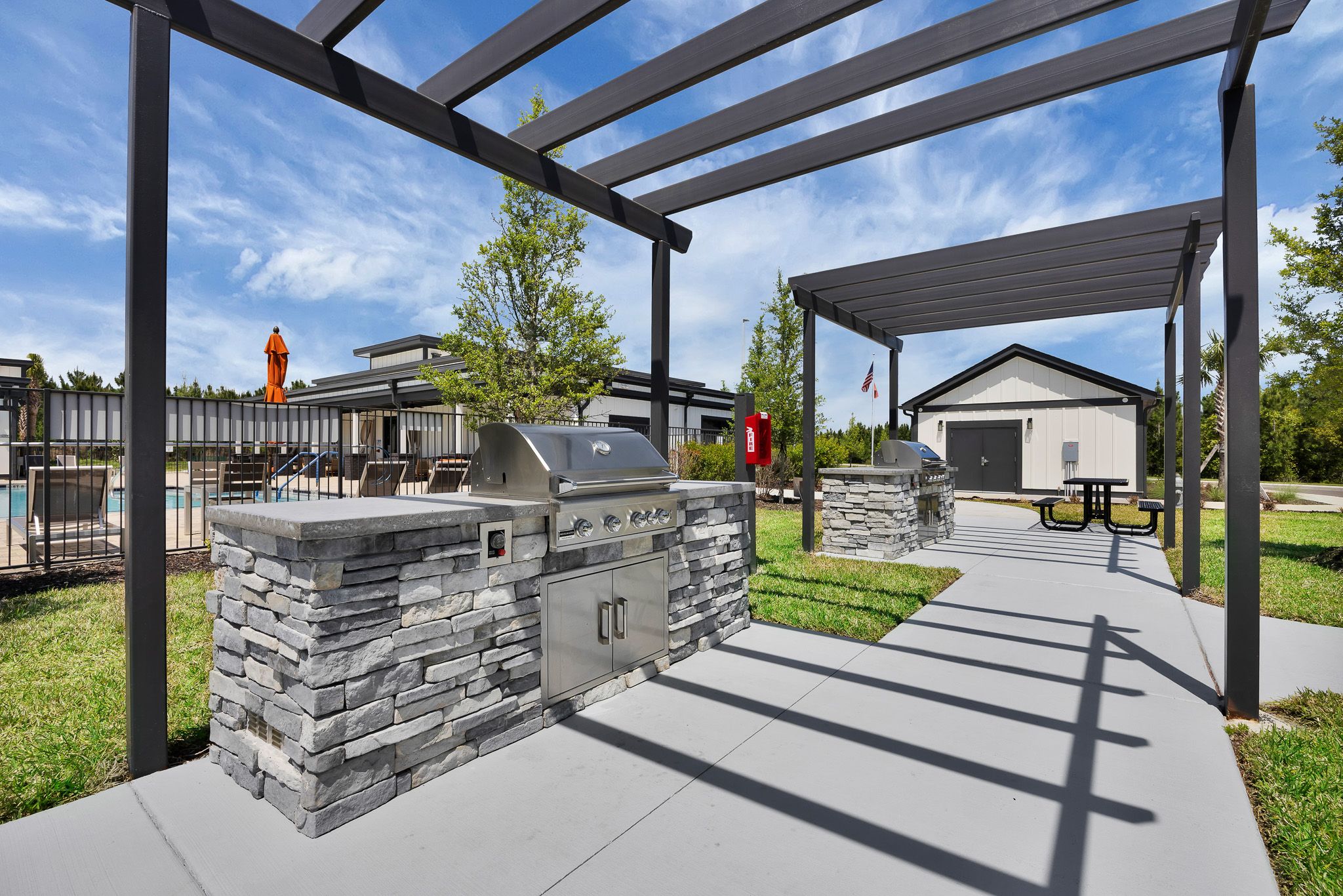 Outdoor grilling area with stone barbecues, shaded pergolas, picnic tables, and a pool in the background.